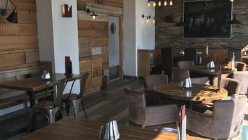 Een modern restaurantinterieur met houten tafels en stoelen, hanglampen en een grote flatscreen televisie aan de muur.