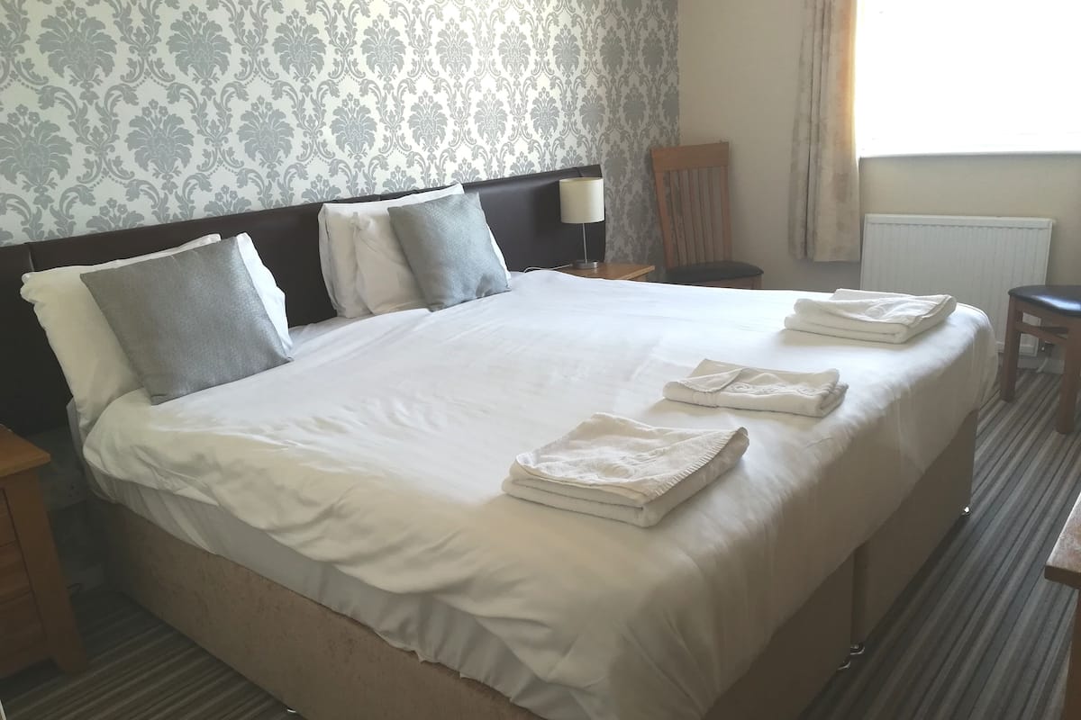 A neatly made bed with white linens and pillows, a wooden nightstand with a lamp, and a chair in a hotel room with patterned wallpaper.