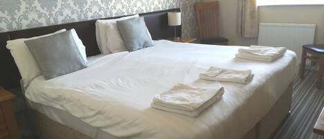 A neatly made bed with white linens and pillows, a wooden nightstand with a lamp, and a chair in a hotel room with patterned wallpaper.