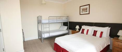 Una habitación de hotel con literas, una cama grande con ropa de cama roja y blanca, una mesita de noche con lámpara y un cuadro enmarcado en la pared.
