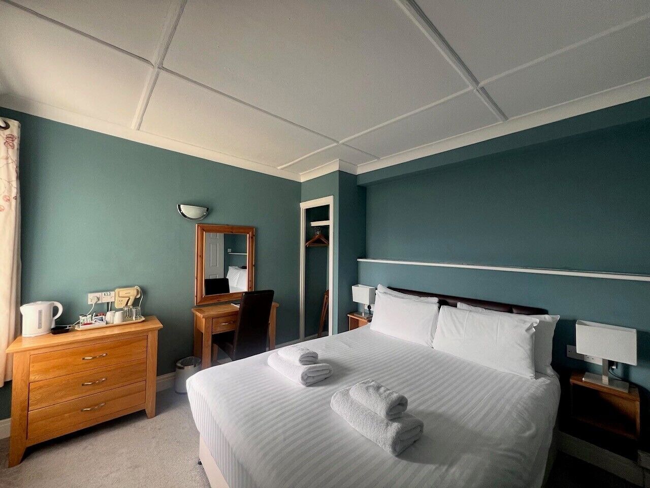 Une chambre d’hôtel avec un grand lit, une commode en bois, un bureau avec une chaise, un miroir et une table de chevet avec une lampe.