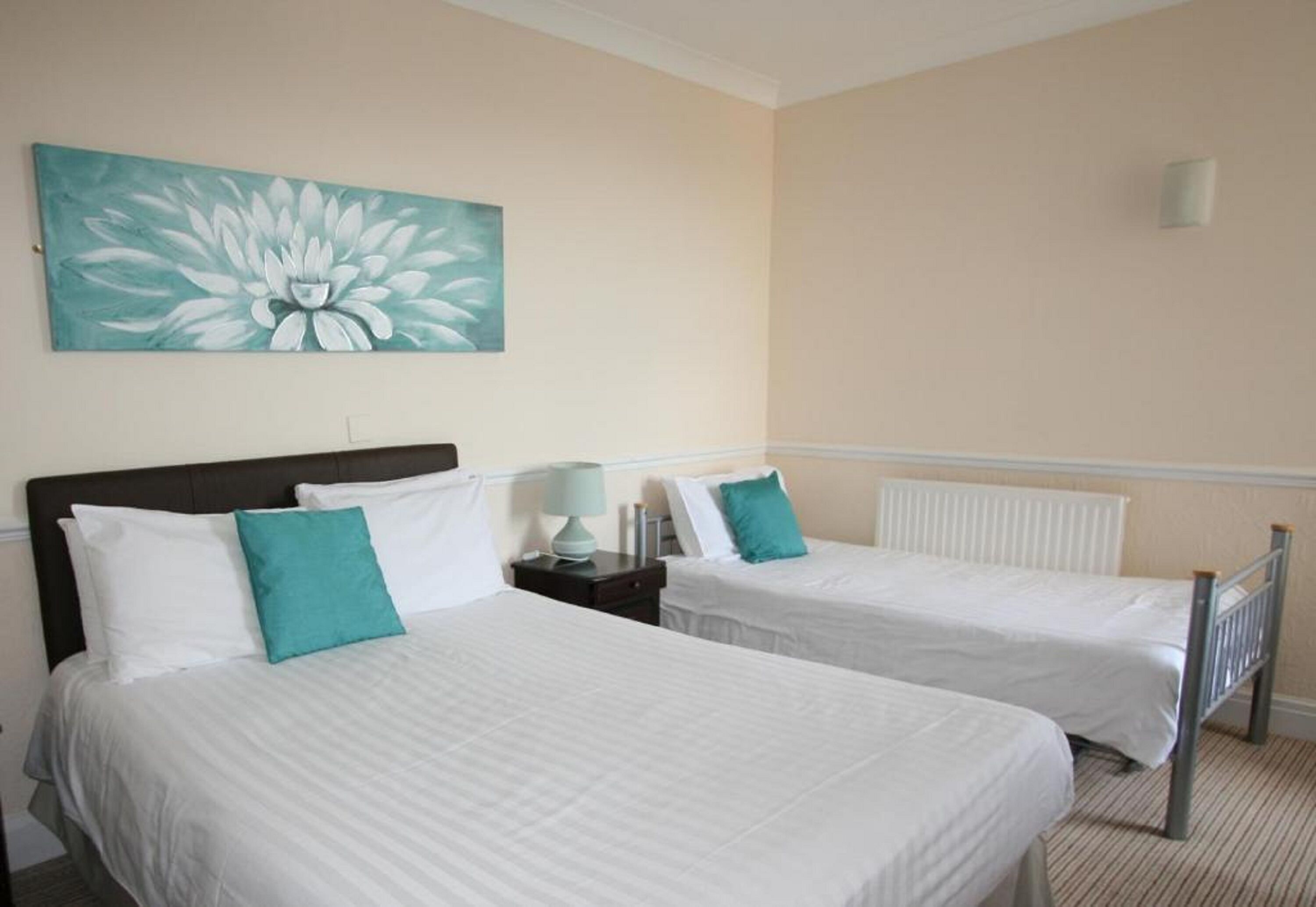 Habitación de hotel con dos camas individuales, ropa de cama blanca, almohadillas color aguamarina, una mesita de noche con lámpara y un cuadro grande con motivos florales en la pared.