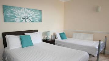 Habitación de hotel con dos camas individuales, ropa de cama blanca, almohadillas color aguamarina, una mesita de noche con lámpara y un cuadro grande con motivos florales en la pared.