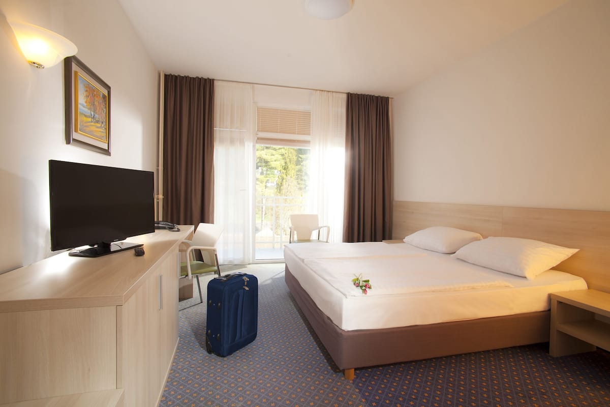A hotel room with a bed, a desk with a television, a suitcase, and a chair.