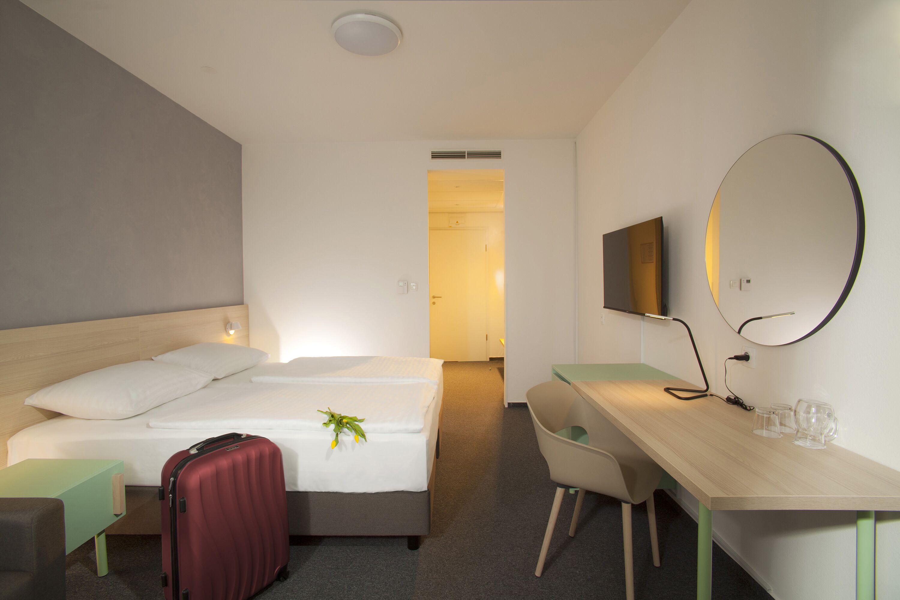 Une chambre d’hôtel avec un lit, un bureau, une chaise, une valise et un miroir.