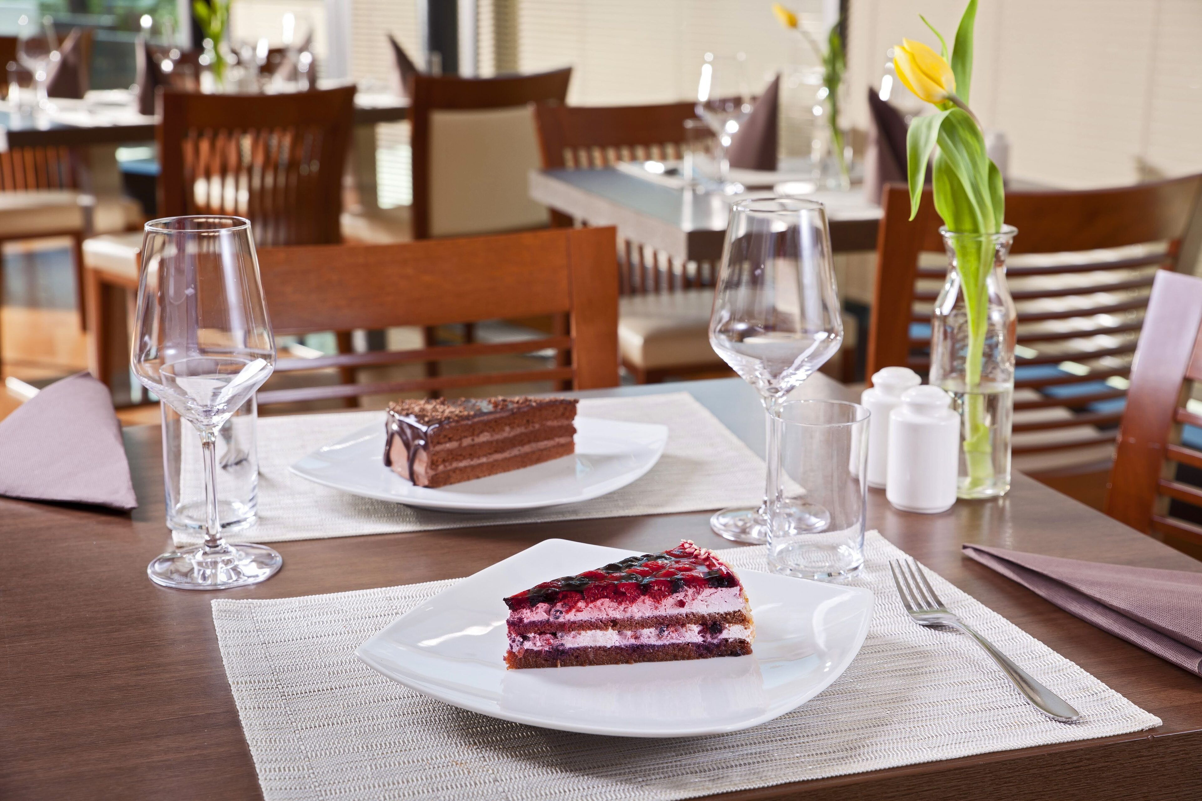 A dining table with a slice of cake, a glass of water, and a vase with a yellow tulip.