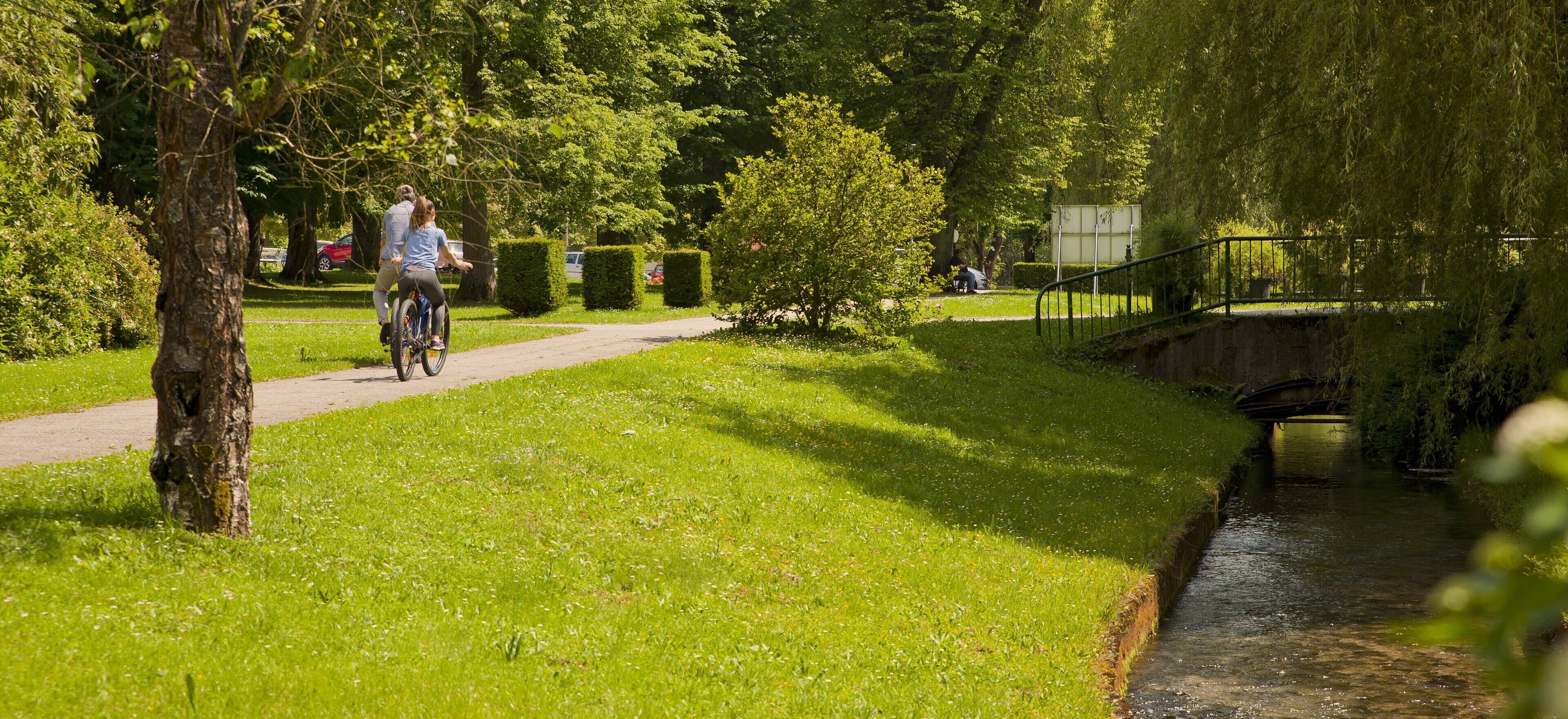 A park pathway with a cyclist, lush greenery, a small bridge, and a water channel.