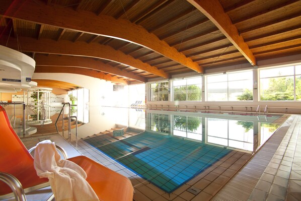 Una piscina interior con vigas de madera, ventanas de vidrio transparente y sillones reclinables naranja.