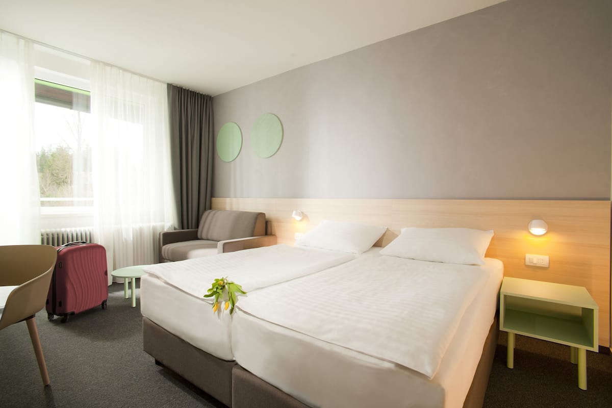 A hotel room with a bed, a chair, a small table, a nightstand, and a window with curtains.