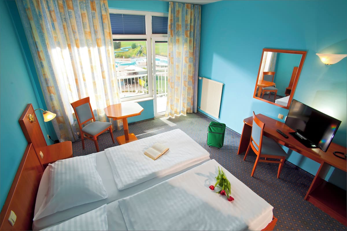 A hotel room with two beds, a desk with a TV, a chair, a small table, and a window with curtains.