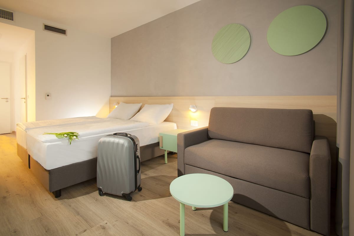 A hotel room with a bed, a sofa, a small table, and a suitcase.