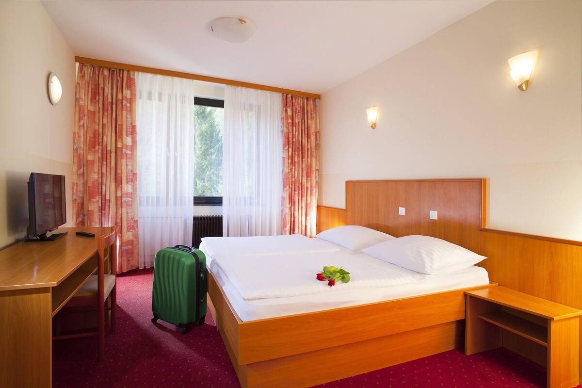 A hotel room with a wooden bed, a desk, a green suitcase, and a television.
