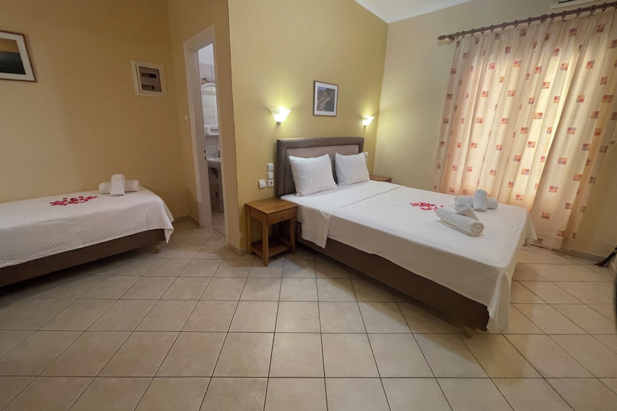 A hotel room with two single beds, a larger bed, a nightstand, and a bathroom visible through an open door.