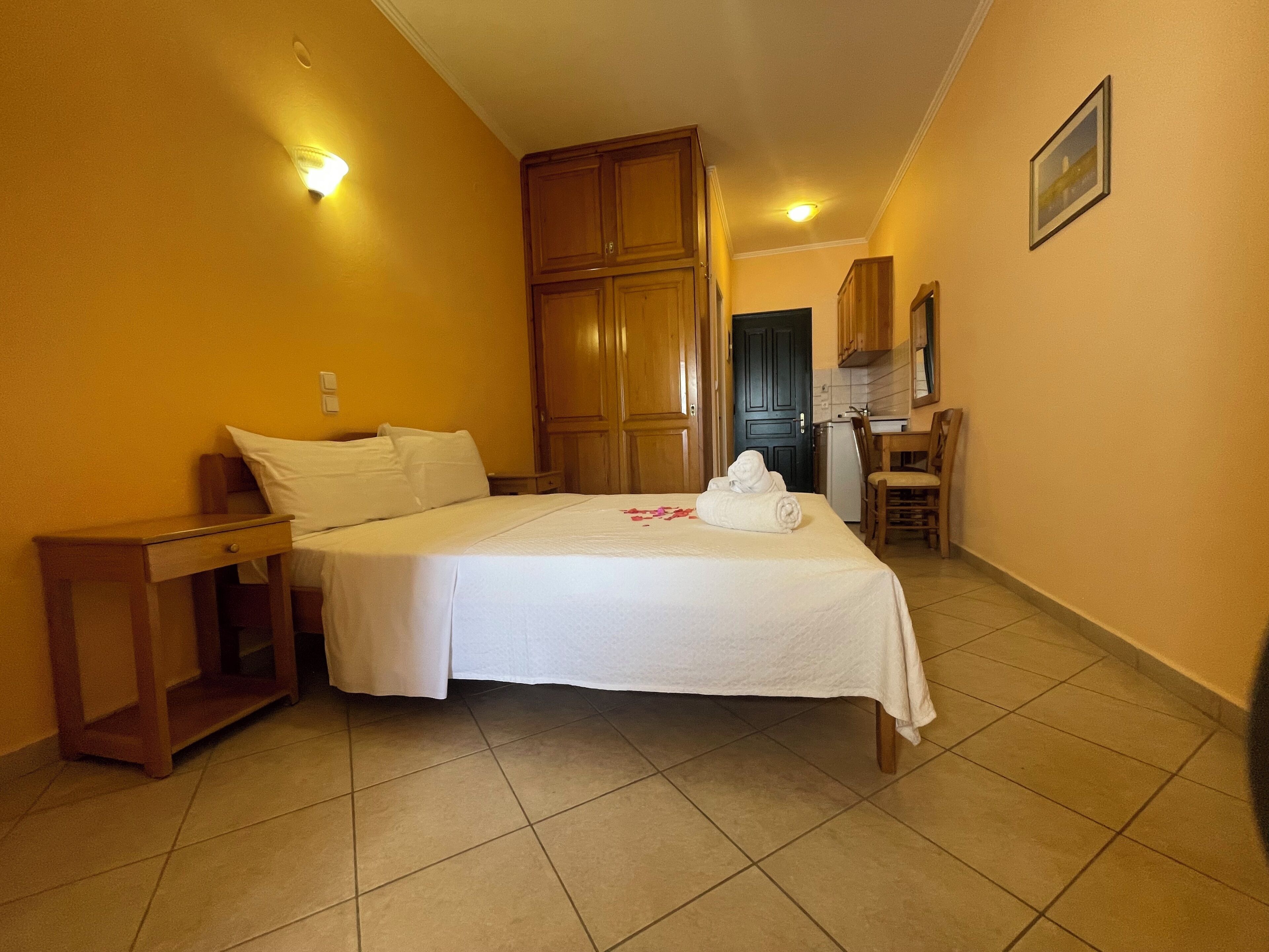A hotel room with a bed, a nightstand, a desk, a chair, a kitchenette, and a door.