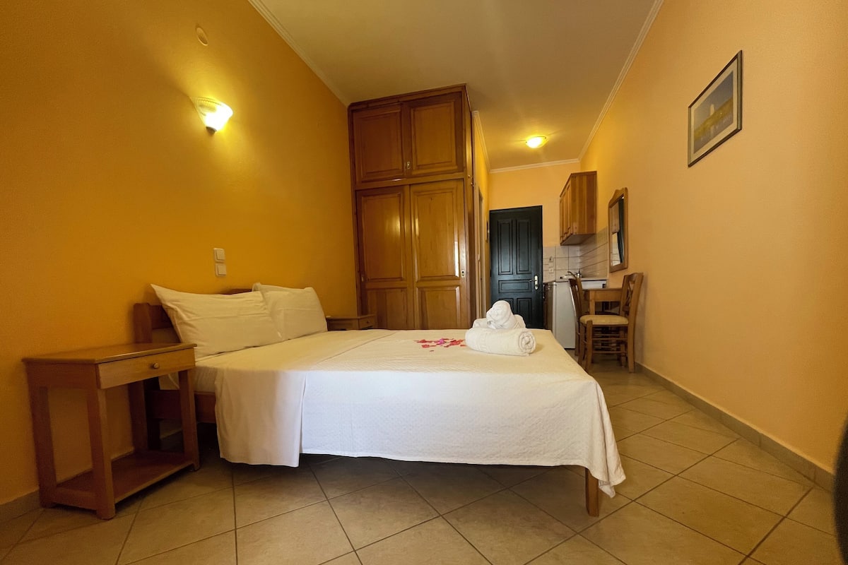 A hotel room with a bed, a nightstand, a desk, a chair, a kitchenette, and a door.