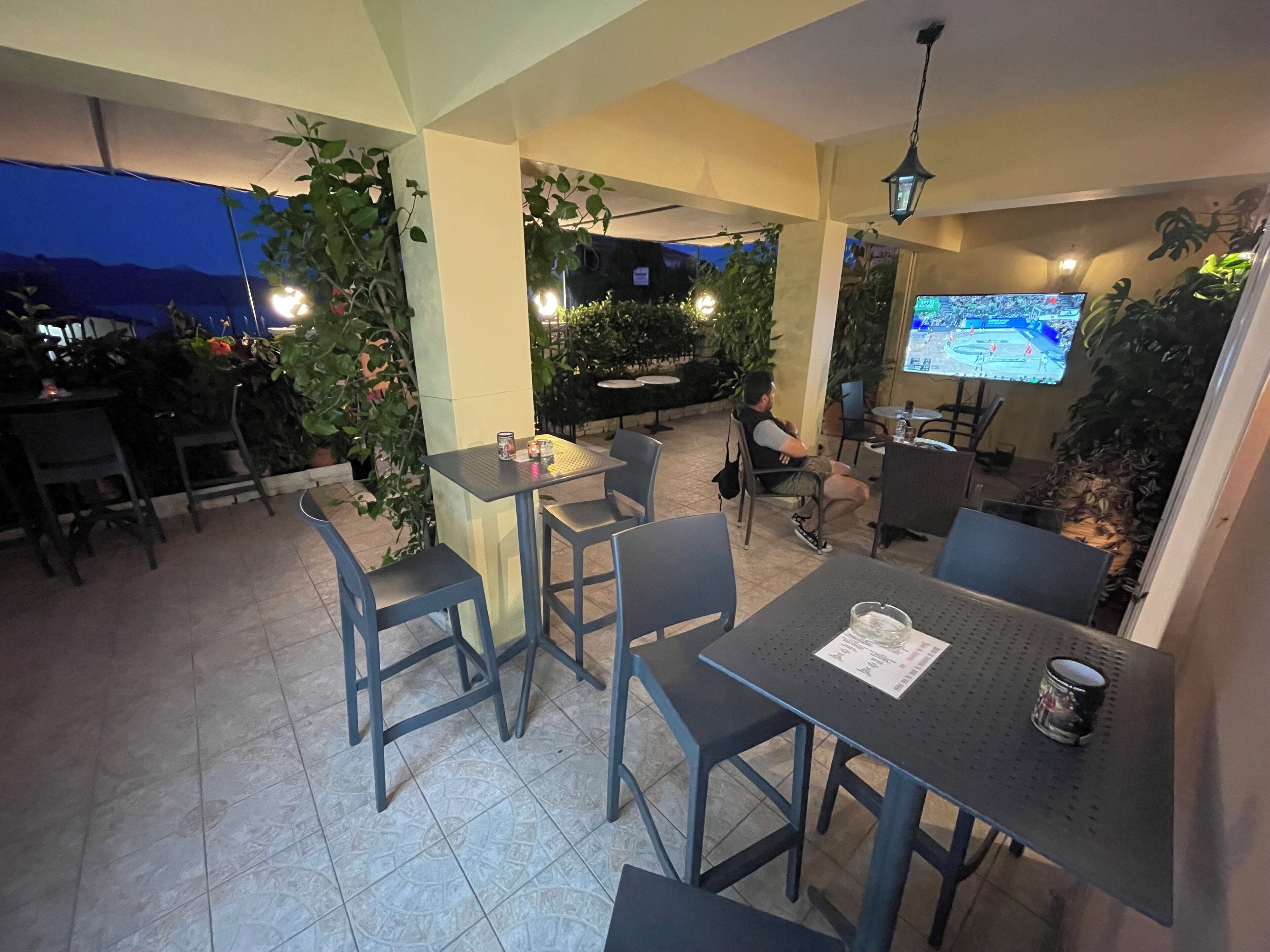 An outdoor patio area with tables and chairs, surrounded by plants and a TV screen displaying a map.