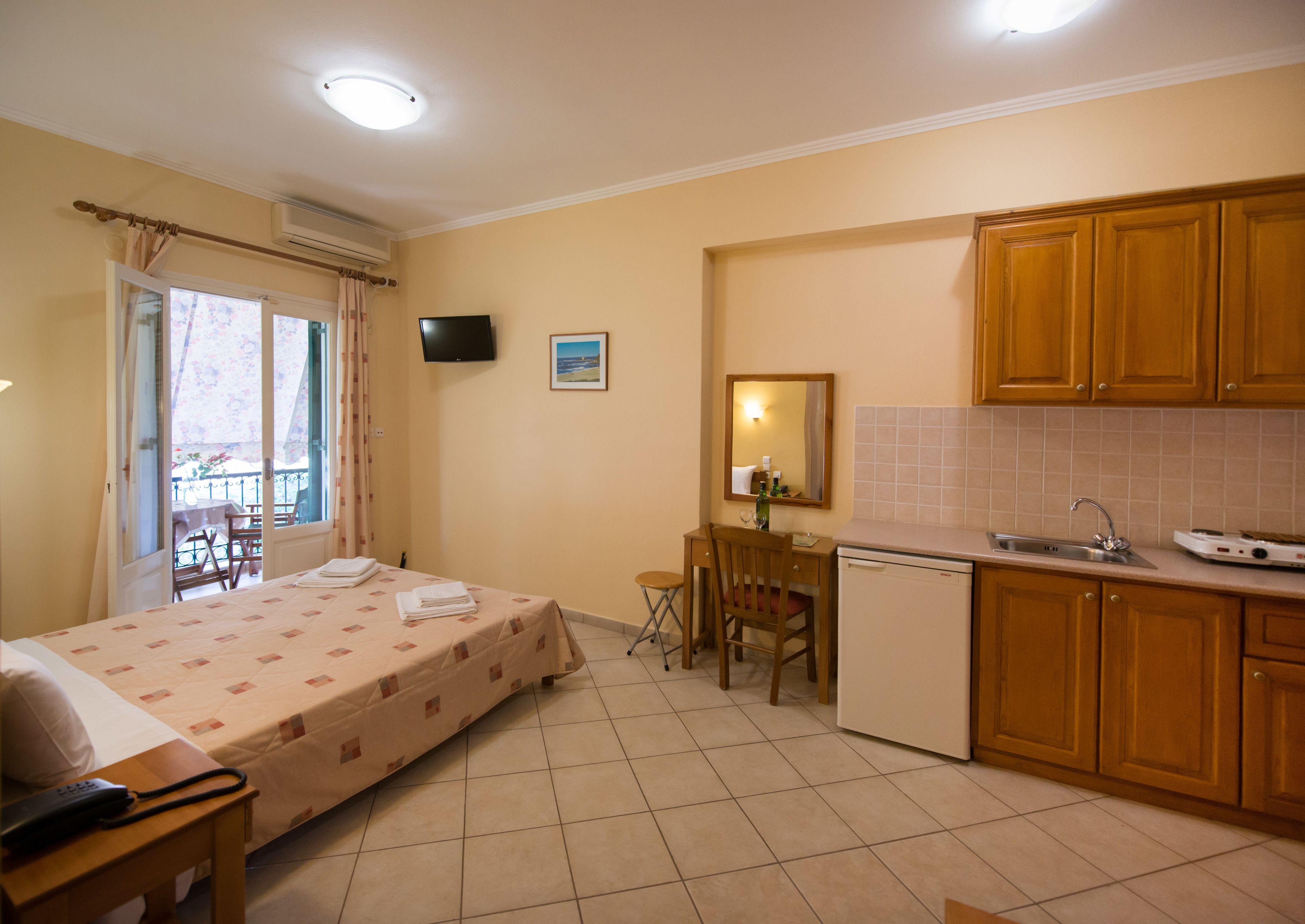 A compact hotel room with a bed, a small desk, a chair, a kitchenette, and a balcony with a table and chairs.