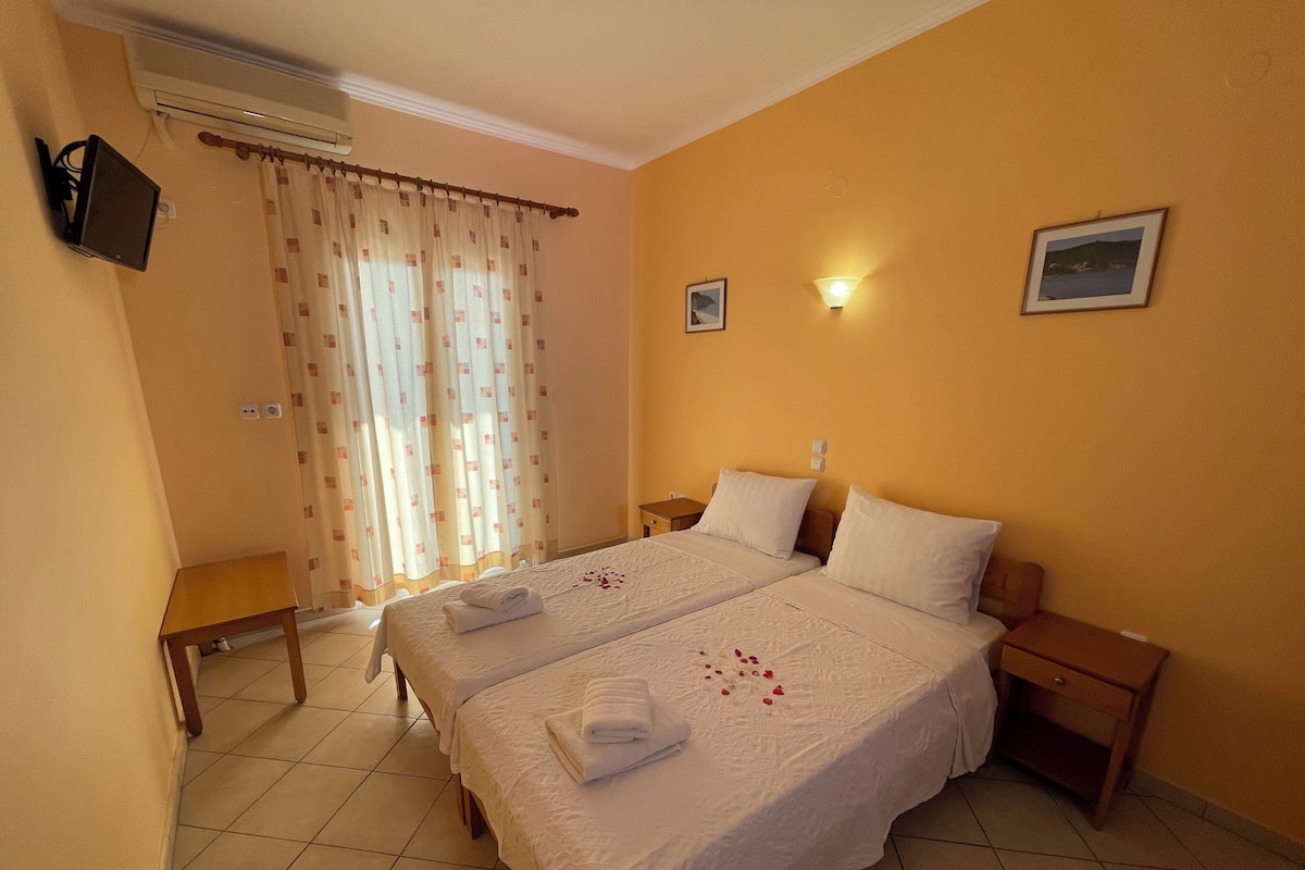 A hotel room with two beds, a small table, a bedside table, a wall-mounted TV, and a window with curtains.