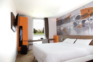 Habitación de hotel con una cama grande, un escritorio con computadora, un perchero y vistas a la ciudad a través de la ventana.