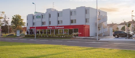 Un edificio moderno con fachada roja y blanca, un acceso cubierto y vista a la calle con coches y vegetación.