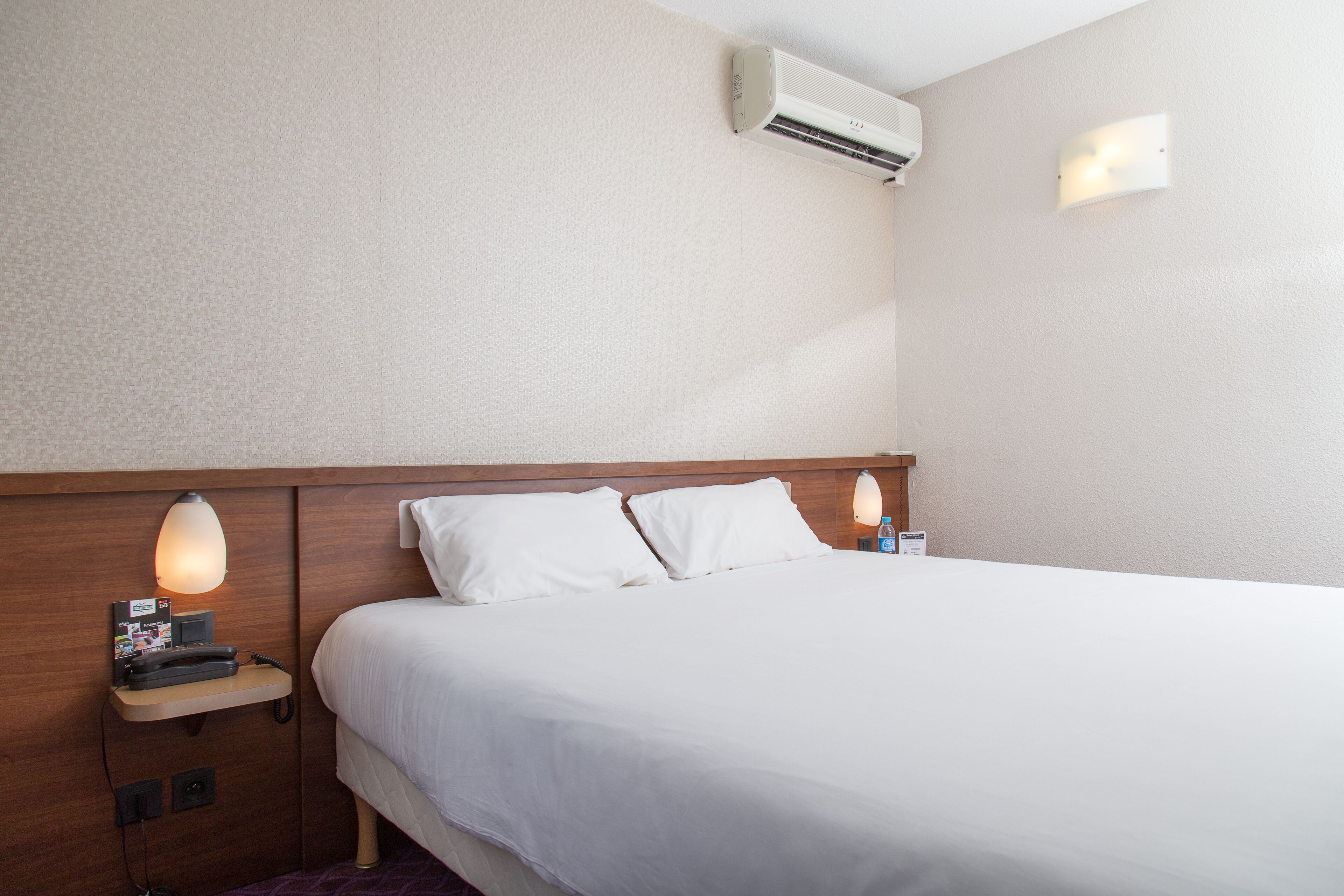 A hotel room with a bed, bedside lamp, air conditioning unit, and a small table with a phone and a brochure.