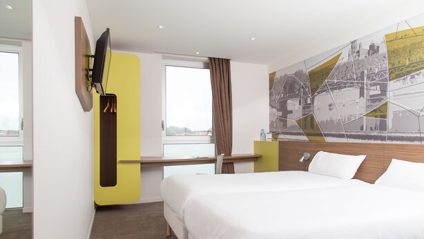 Een moderne hotelkamer met een groot bed, een bureau met een flatscreen-tv, een groot raam en een stadsmuur op de muur.