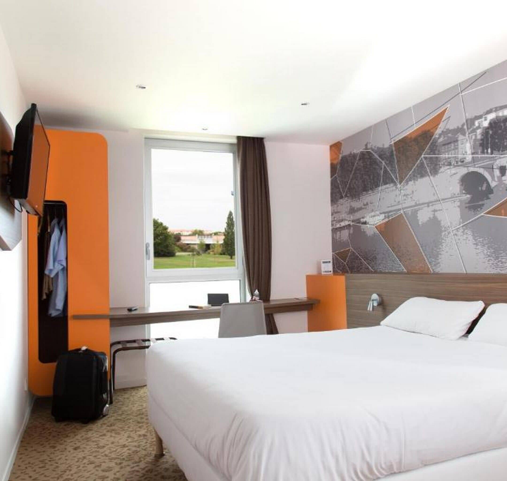 A hotel room with a bed, a desk, a chair, a TV, and a large window with a view of greenery.