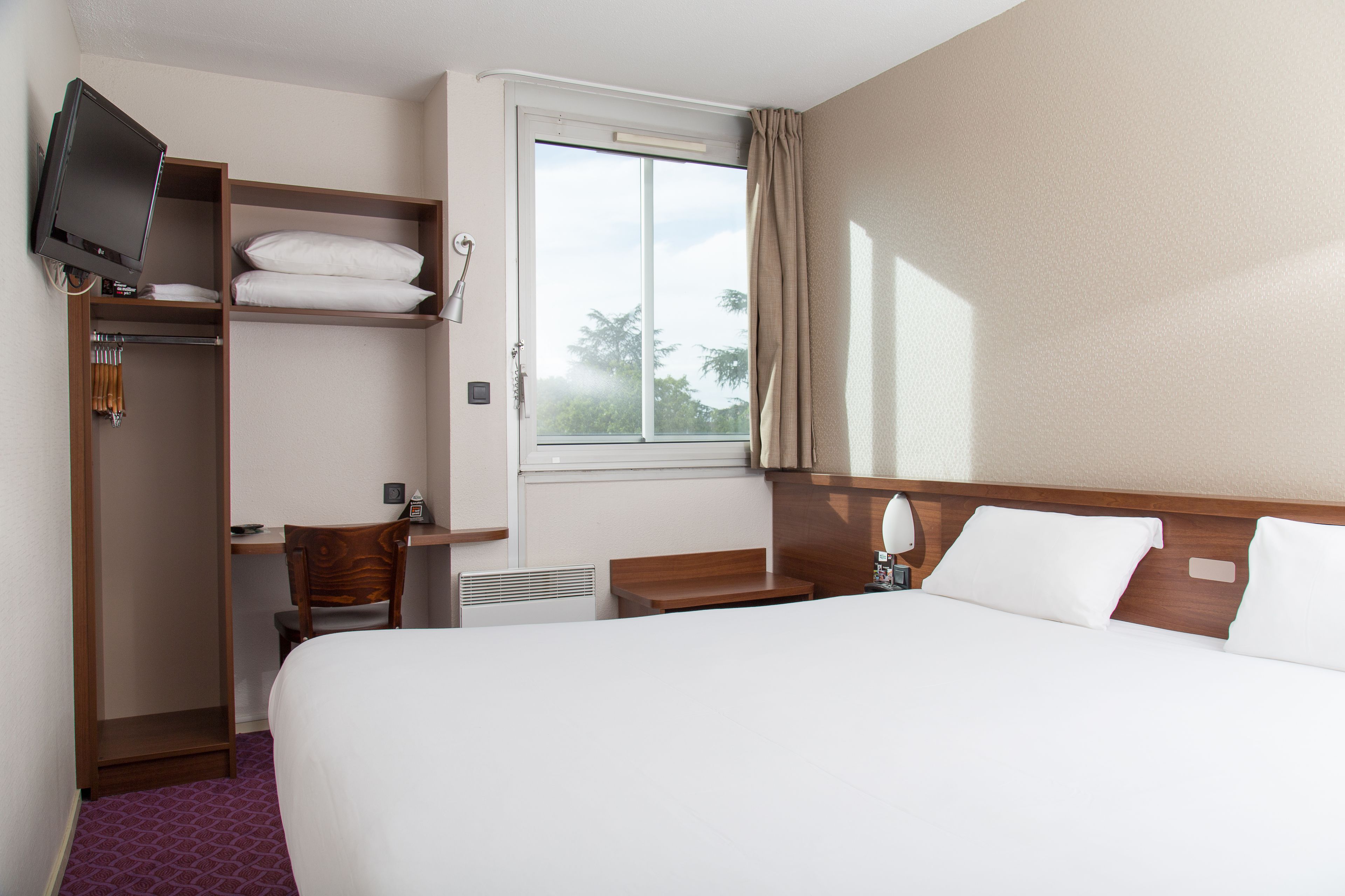 Una habitación de hotel con cama, escritorio, silla, televisor, ventana y armario.