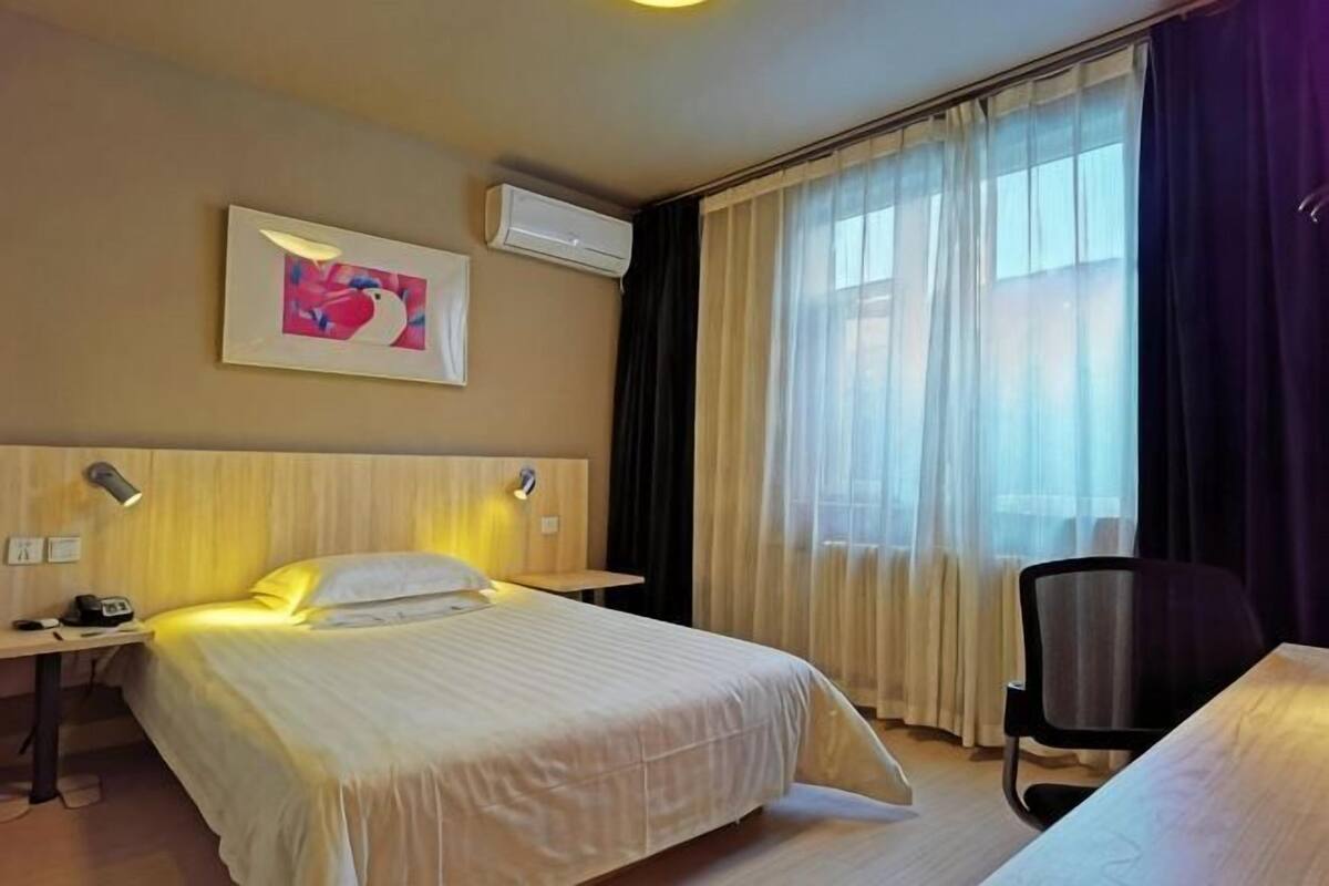 A hotel room with a bed, bedside tables, a desk, a chair, an air conditioner, and a window with curtains.