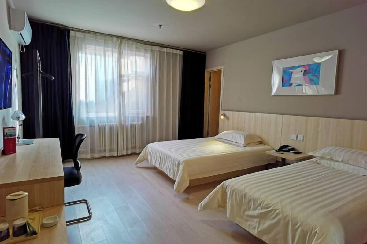 A hotel room with two beds, a desk with a chair, a window with curtains, and a painting on the wall.