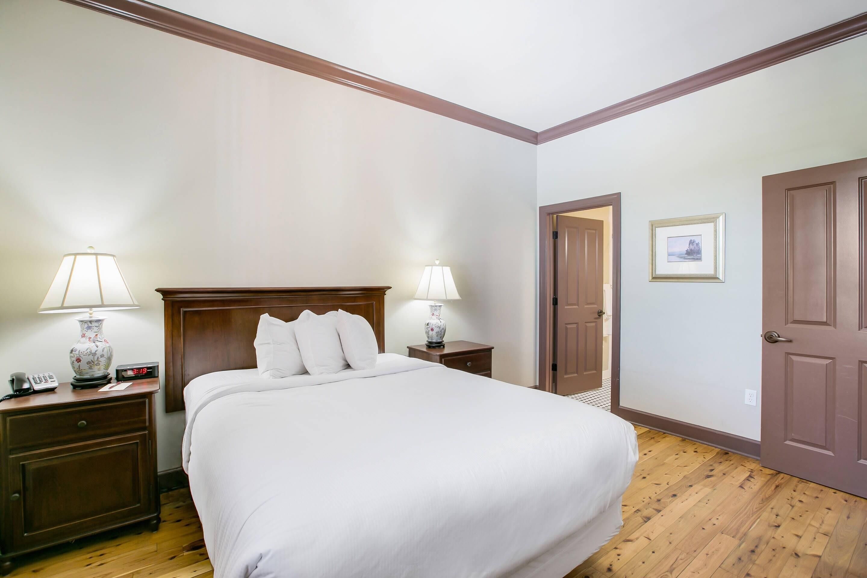 A hotel room with a large bed, two bedside tables with lamps, a wooden headboard, a door, and a framed picture on the wall.