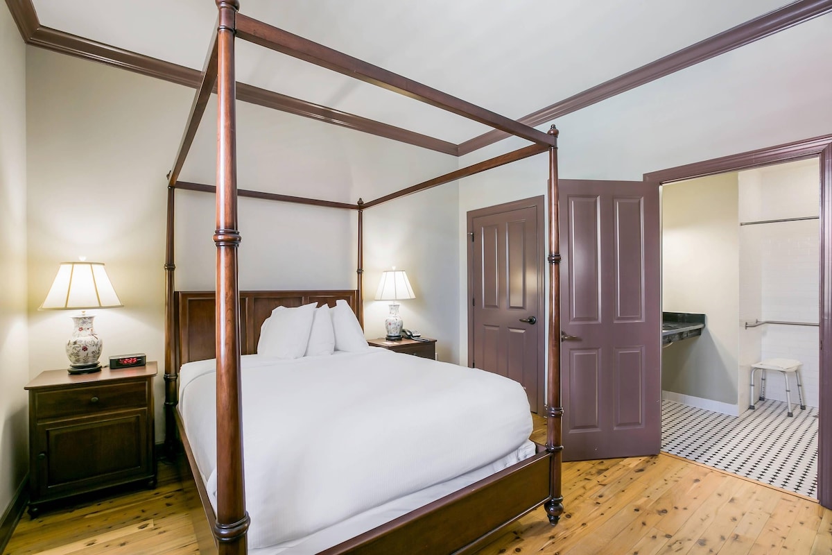 A four-poster bed with a wooden frame, two bedside tables with lamps, a door, and a small stool.