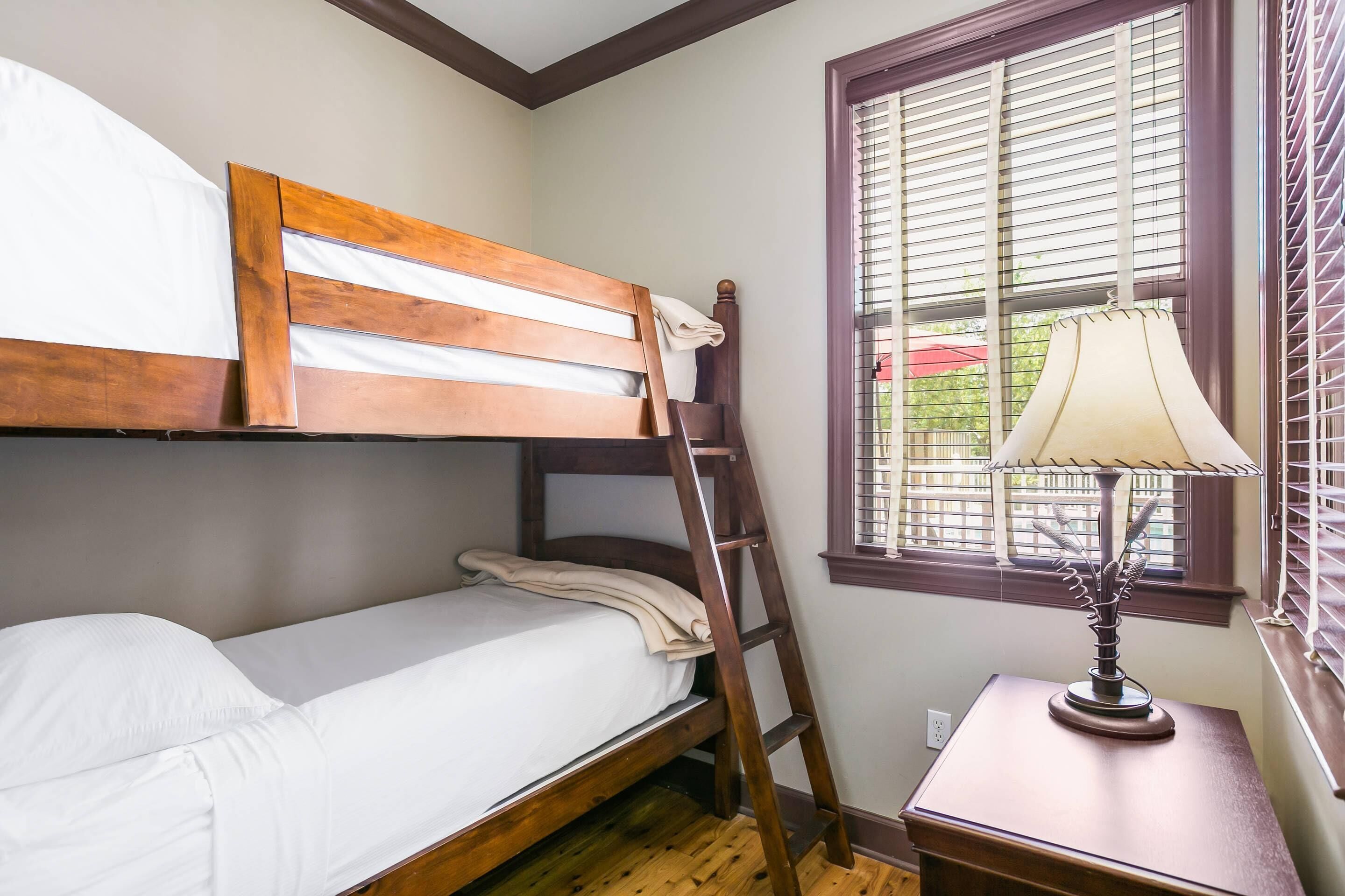 A bunk bed with a wooden headboard, a bedside table with a lamp, and a window with blinds.
