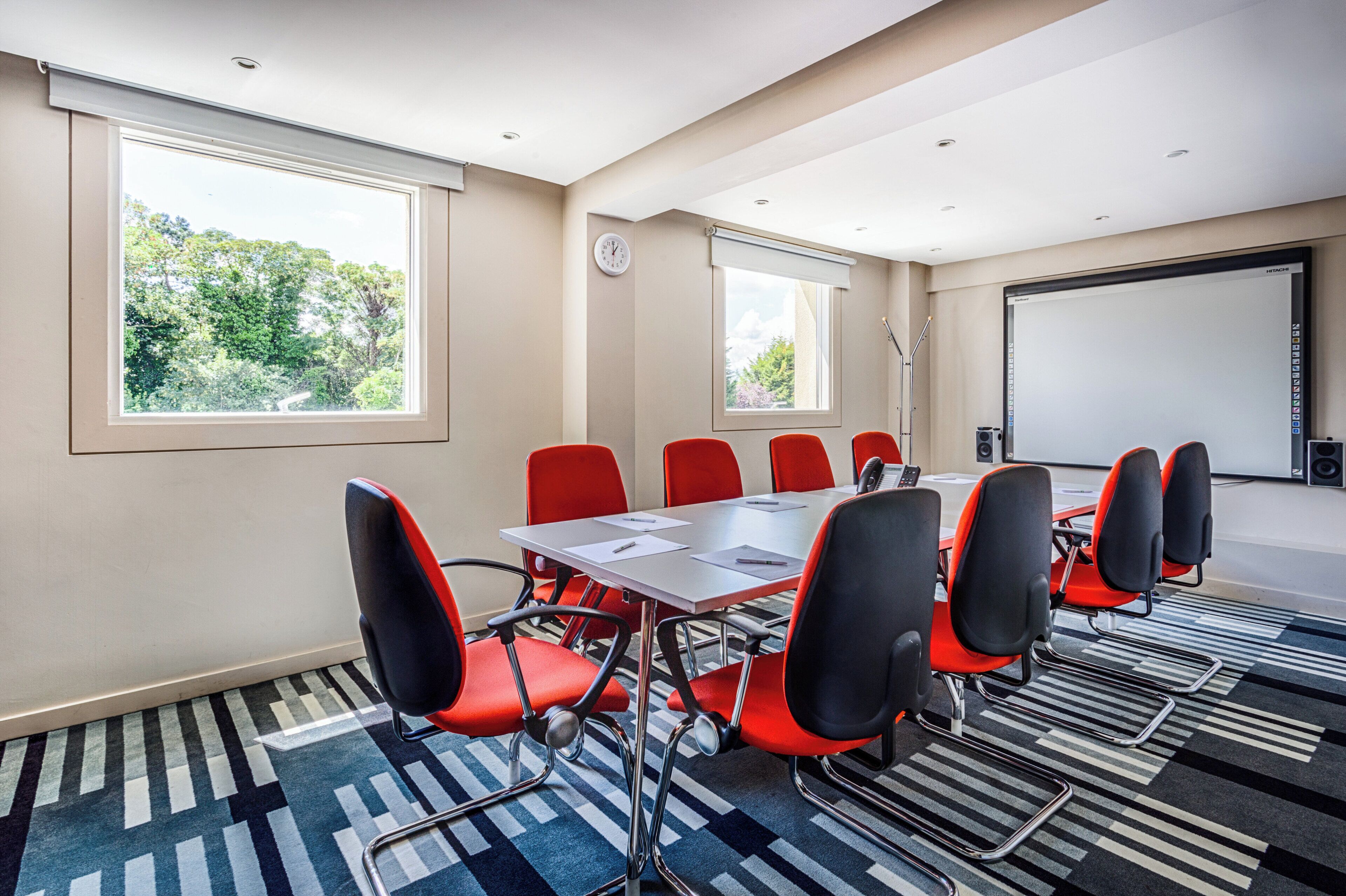 Sala de conferências com uma mesa grande, cadeiras vermelhas, tela de projetor e um relógio na parede.