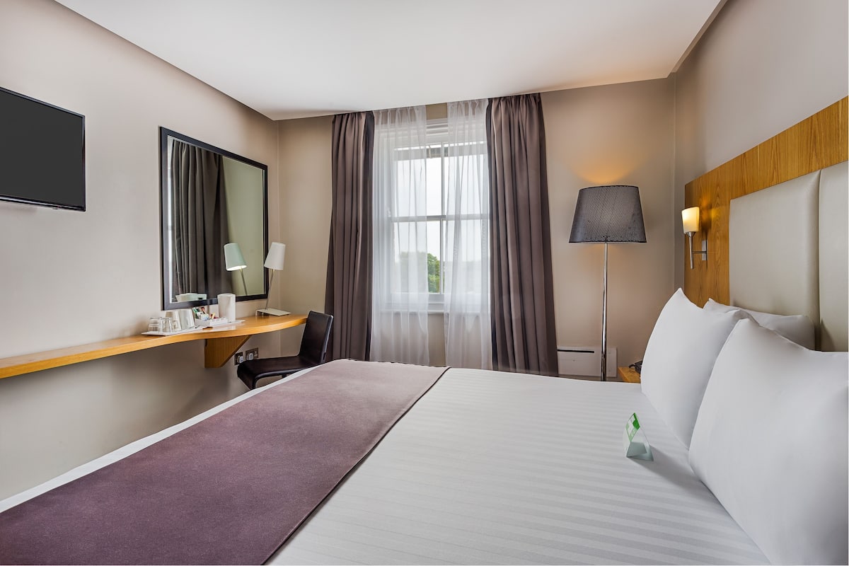 A hotel room with a large bed, a desk with a chair, a flat-screen TV, a lamp, and a window with curtains.