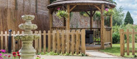A wooden gazebo with a thatched roof, surrounded by a wooden fence, and a stone fountain in the foreground.