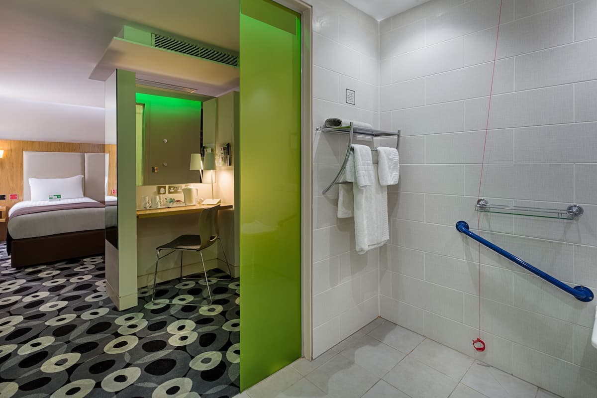 A hotel room with a bed, a desk, a chair, a shower area with a grab bar, and a patterned floor.