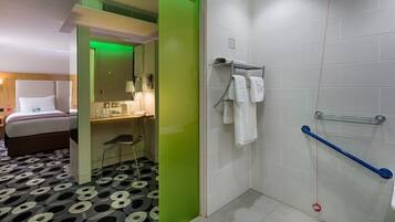 A hotel room with a bed, a desk, a chair, a shower area with a grab bar, and a patterned floor.