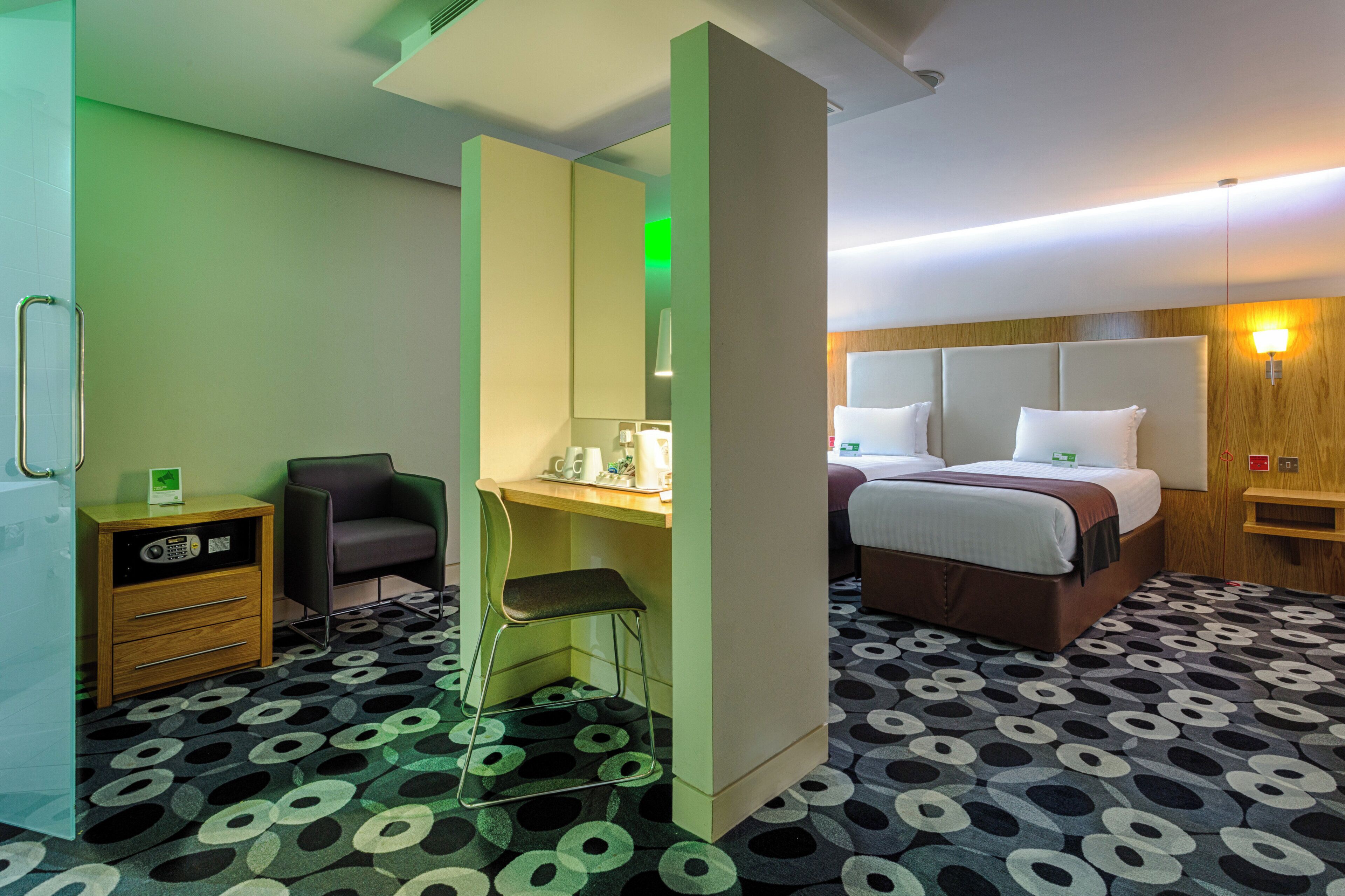 A hotel room with a bed, a desk, a chair, a small cabinet, and a patterned carpet.