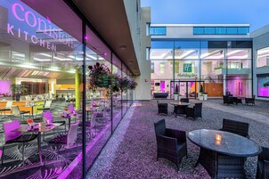 An outdoor seating area with tables and chairs, a glass wall with a restaurant sign, and a Holiday Inn building in the background.