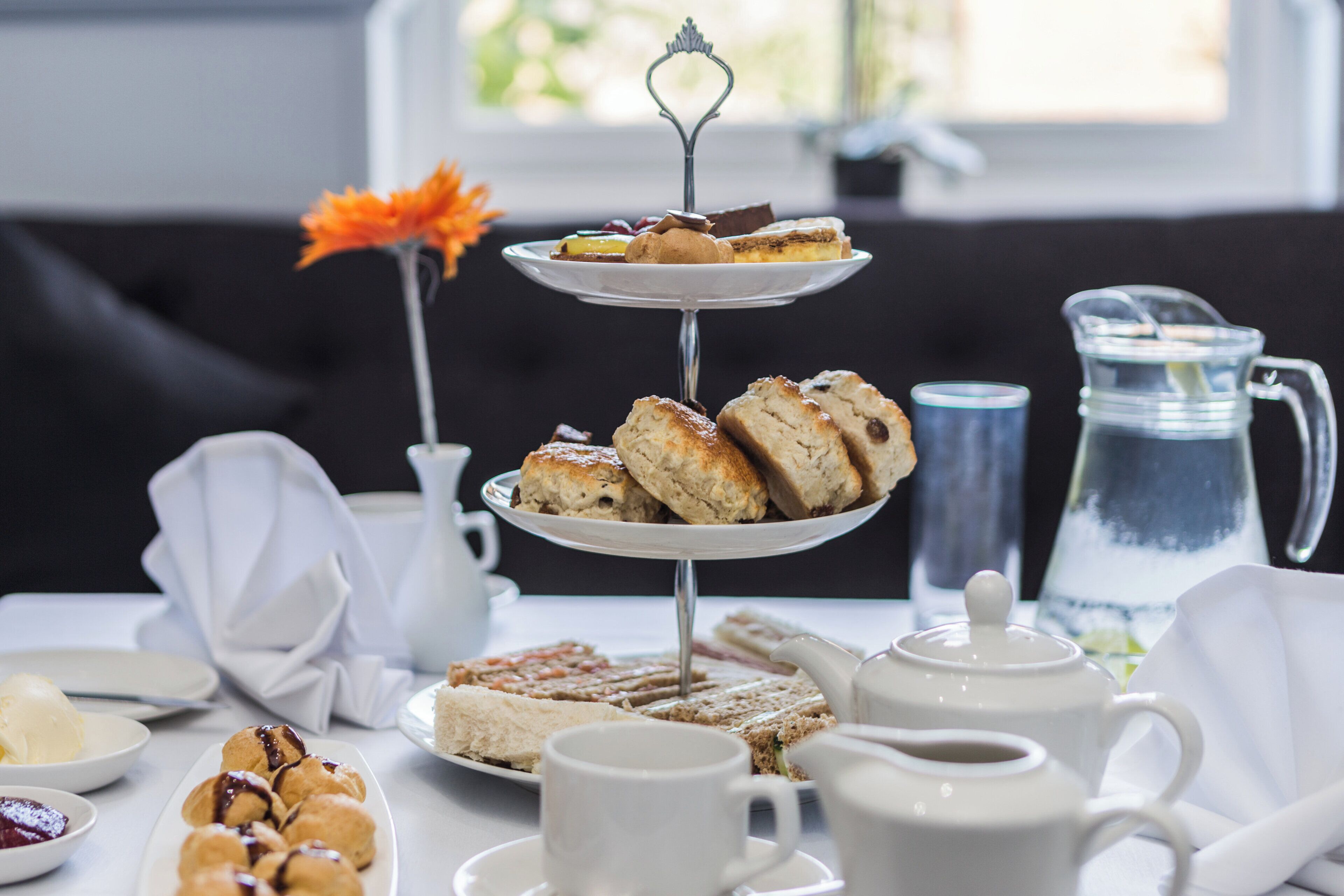 A tiered dessert stand with pastries and a flower, a pitcher, cups, and a teapot on a table.