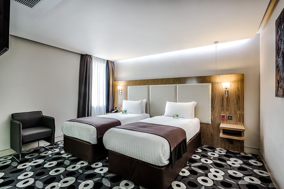 A hotel room with two beds, a chair, a wooden headboard, a patterned carpet, and a nightstand with a lamp.