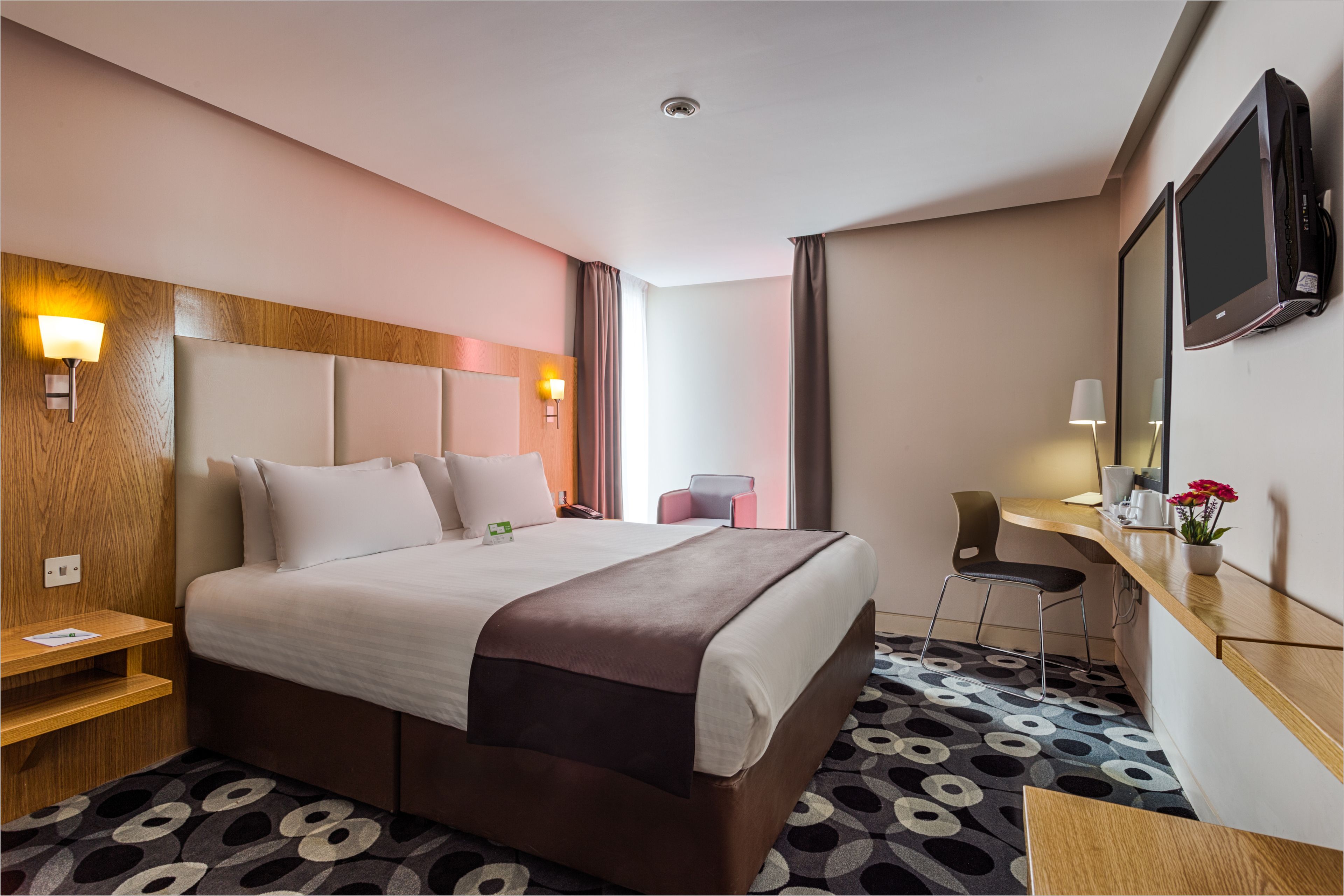 A hotel room with a large bed, a desk with a chair, a TV, and a small table with a flower vase.