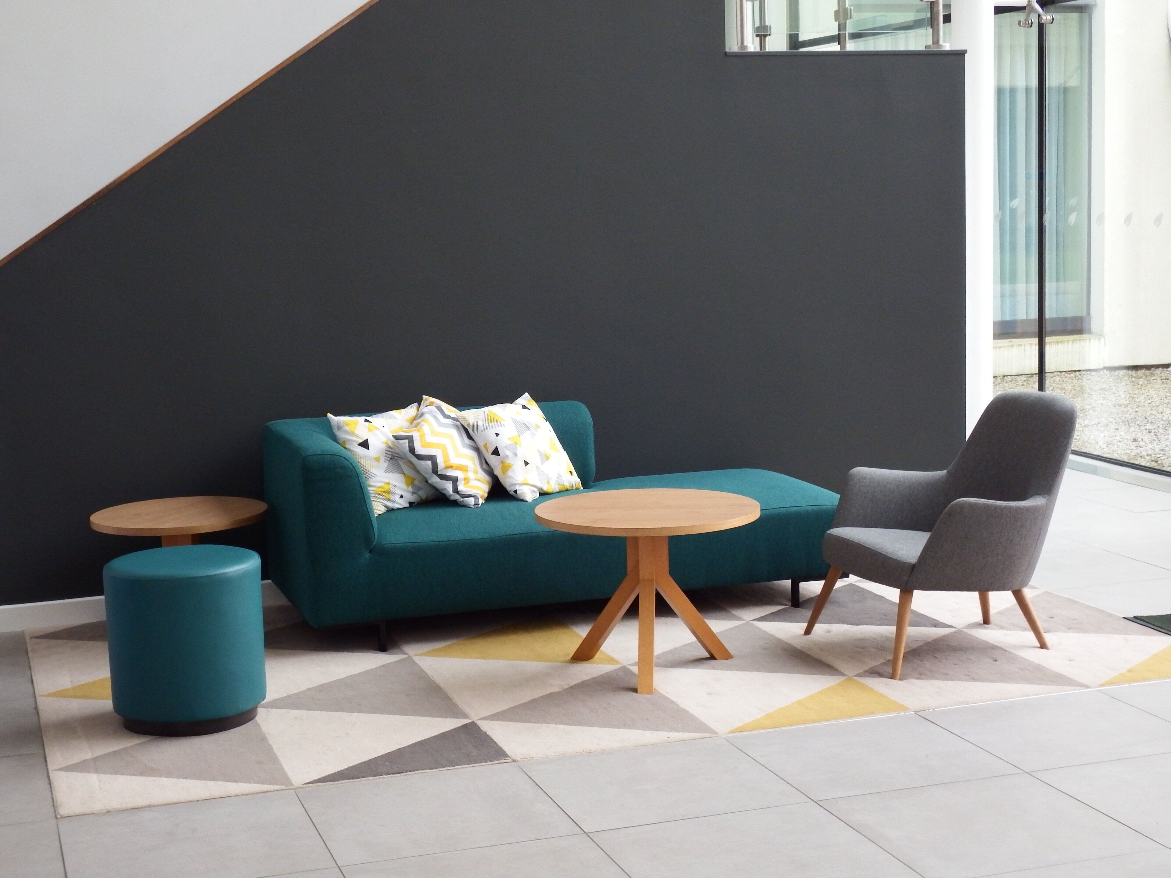A modern lounge area with a teal sofa, a grey armchair, and two round wooden tables.