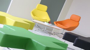 Een moderne kantoorruimte met een gele en oranje stoel, een witte tafel met een schaal fruit, en groene en zwarte stoelen.