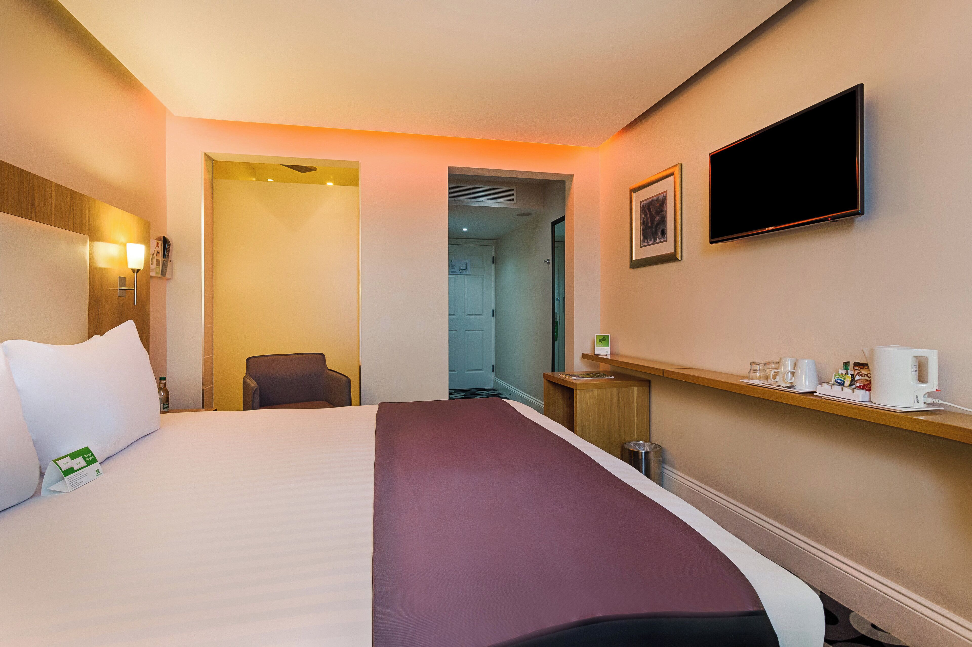 A hotel room with a bed, a chair, a desk with a coffee maker, a television, and a door leading to another room.