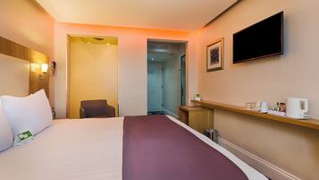 A hotel room with a bed, a chair, a desk with a coffee maker, a television, and a door leading to another room.