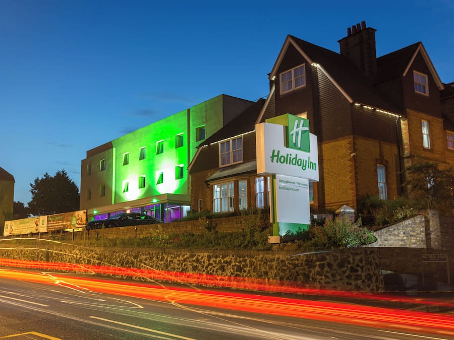 Um hotel Holiday Inn com fachada iluminada em verde, uma parede de pedra e uma placa de identificação.