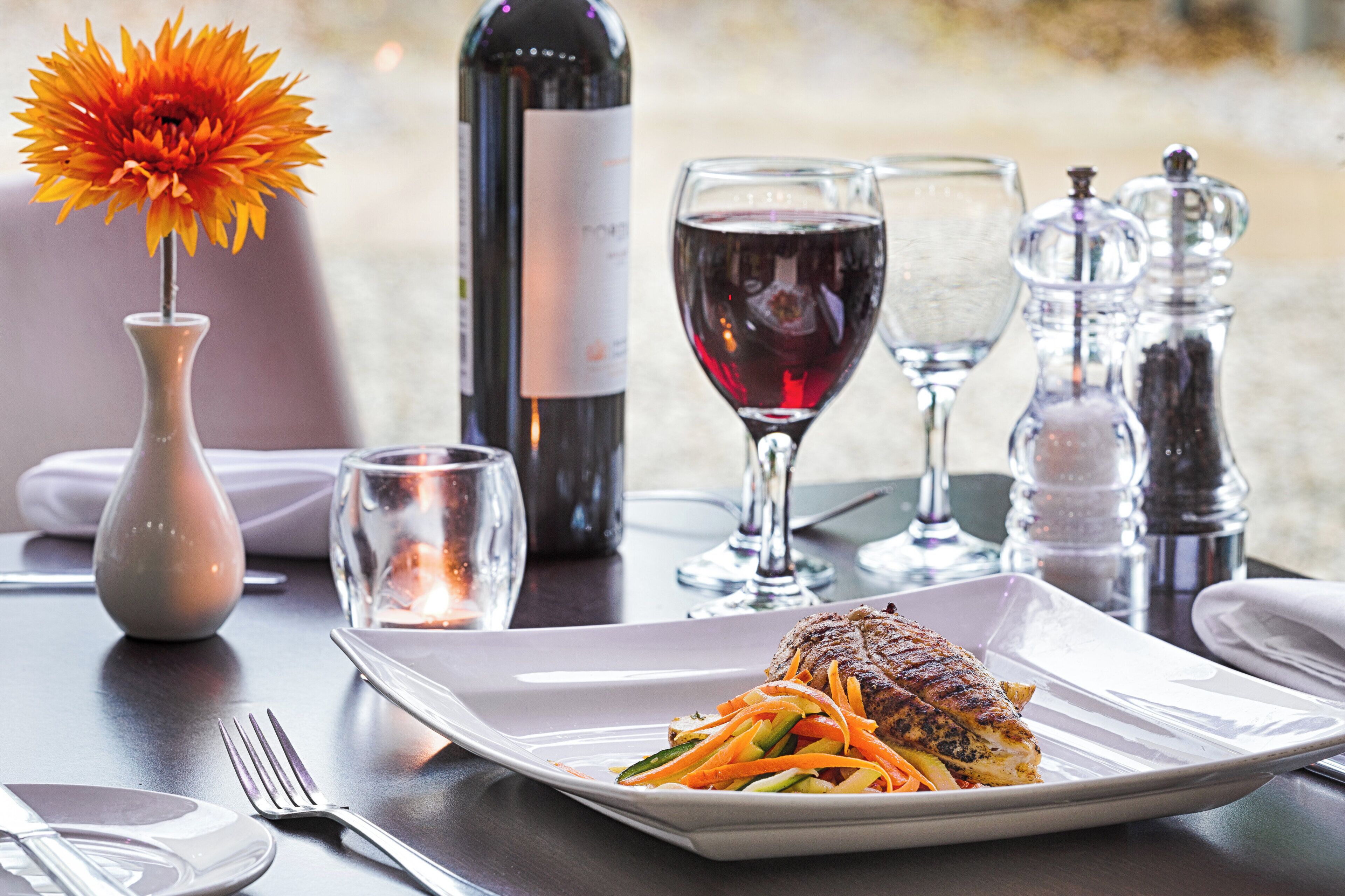 A dining table with a plate of food, wine glasses, a bottle of wine, and a vase with a flower.