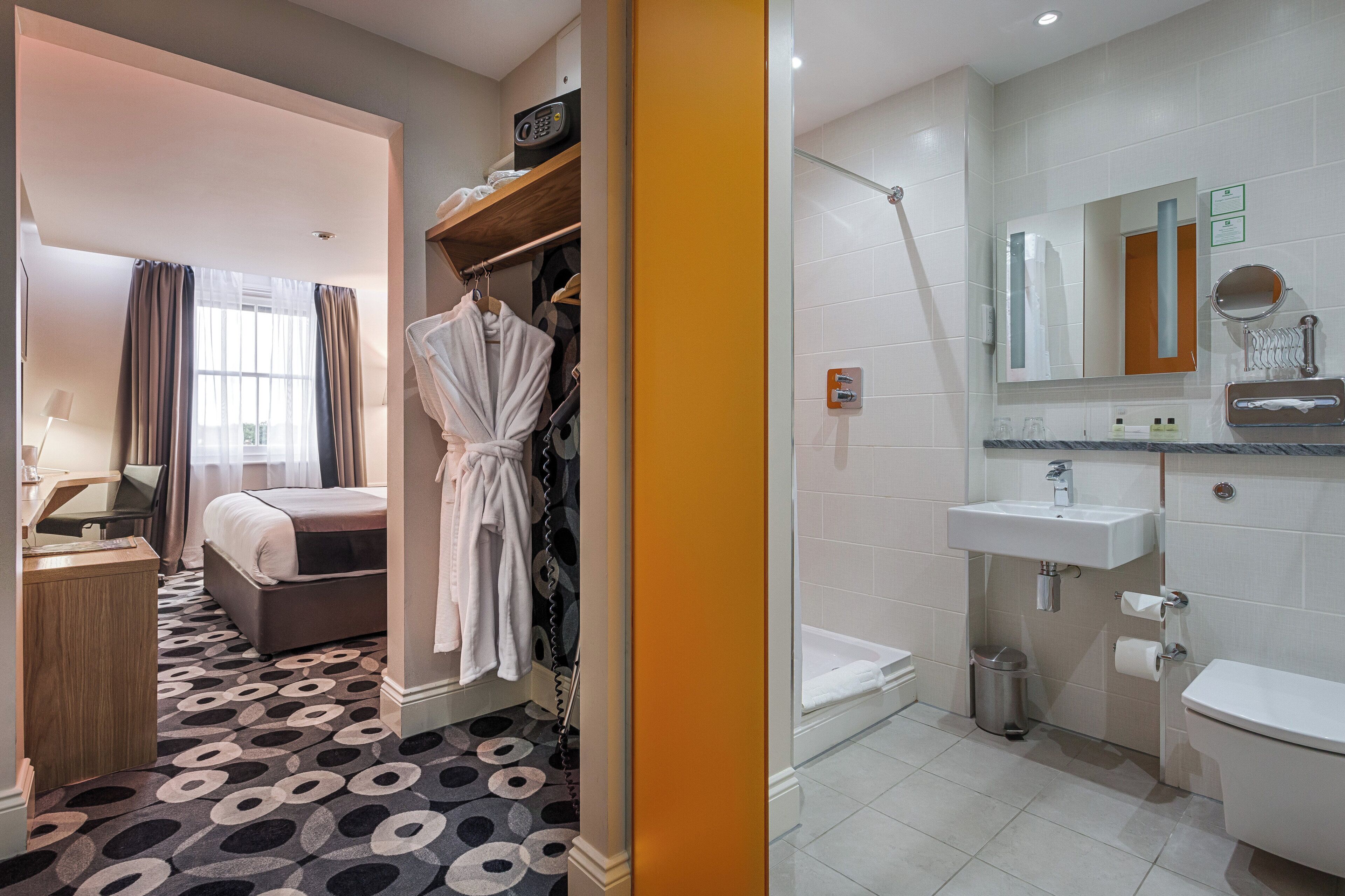 Quarto de hotel com cama, escrivaninha, roupão e banheiro com pia, espelho e vaso sanitário.